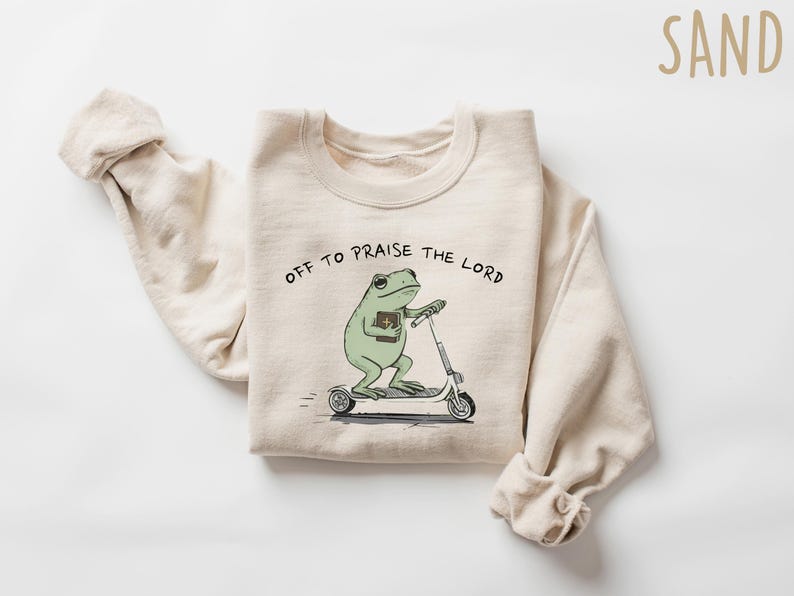 May include: Sand-colored sweatshirt with the text "OFF TO PRAISE THE LORD." The graphic features a green frog riding a scooter while holding a book. The sweatshirt has long sleeves.