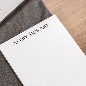Puede incluir: Un bloc de notas blanco con el texto "AVERY STEWART" impreso en negro en la parte superior. El bloc de notas está sobre una superficie gris con un portátil y una planta en maceta en el fondo.