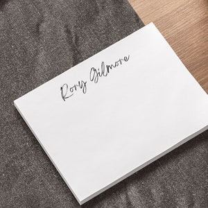 Może przedstawiać: Biały notes z napisem "Rory Gilmore" napisanym czarnym atramentem u góry. Notes leży na szarej, teksturowanej powierzchni.