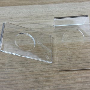 May include: Two clear acrylic display stands, each featuring a rectangular base with a bent support edge and a circular cutout in the center. These transparent holders are designed to support items at an angle, showcasing a smooth, polished finish.