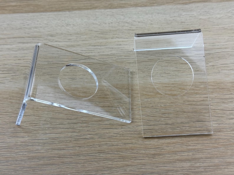 May include: Two clear acrylic display stands, each featuring a rectangular base with a bent support edge and a circular cutout in the center. These transparent holders are designed to support items at an angle, showcasing a smooth, polished finish.