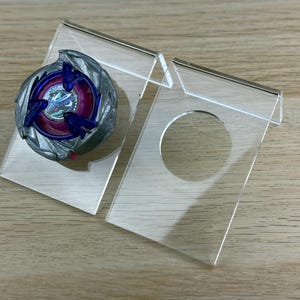 May include: A circular spinning top toy with a metallic silver outer ring, a deep purple center, and blue accents, displayed on a clear acrylic stand. Next to it is an identical empty clear acrylic stand featuring a circular cutout for display.