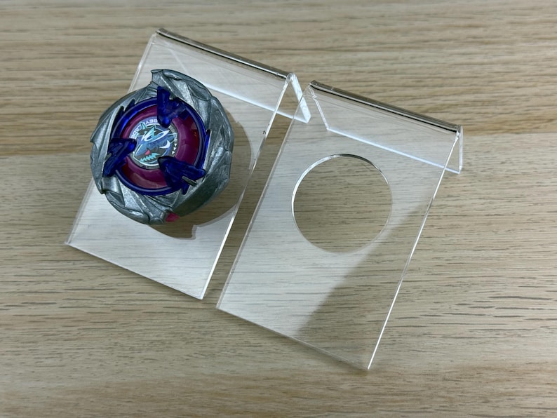 May include: A circular spinning top toy with a metallic silver outer ring, a deep purple center, and blue accents, displayed on a clear acrylic stand. Next to it is an identical empty clear acrylic stand featuring a circular cutout for display.