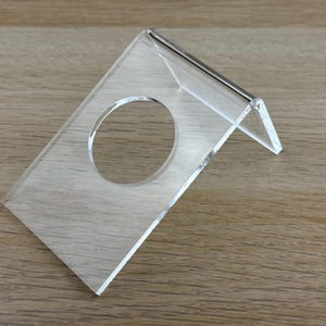 May include: A clear acrylic display stand featuring an angled design with a circular cutout in the center. The stand is constructed from a single piece of transparent material bent to create a stable base and a slanted surface for holding items.