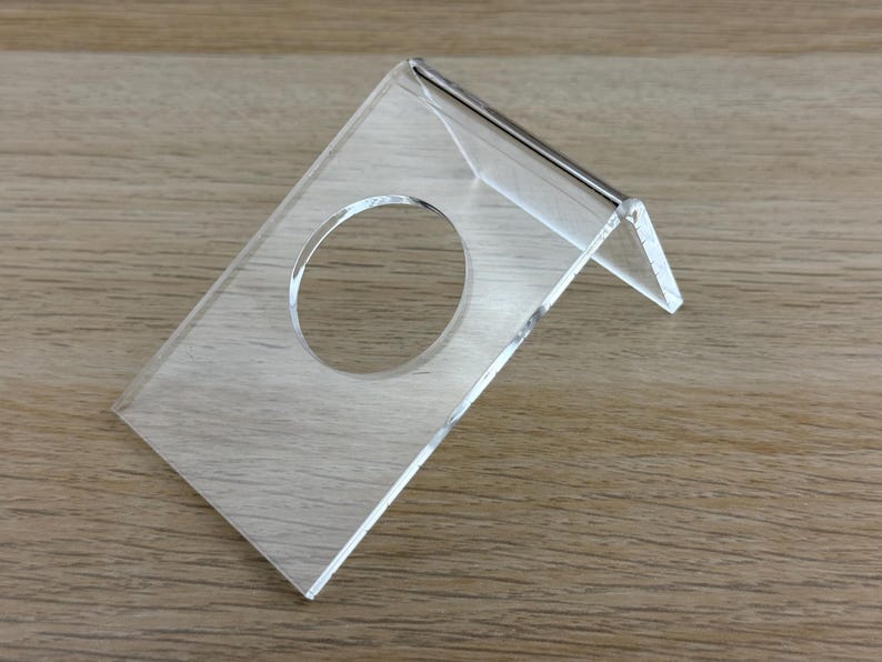 May include: A clear acrylic display stand featuring an angled design with a circular cutout in the center. The stand is constructed from a single piece of transparent material bent to create a stable base and a slanted surface for holding items.