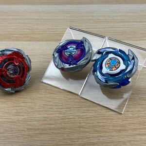 May include: Three circular spinning top toys with metallic silver outer rings. One top is red with a dark center, one is purple with a blue center, and one is blue with a white center featuring a graphic of a blue creature with orange flames.