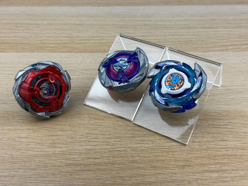May include: Three circular spinning top toys with metallic silver outer rings. One top is red with a dark center, one is purple with a blue center, and one is blue with a white center featuring a graphic of a blue creature with orange flames.