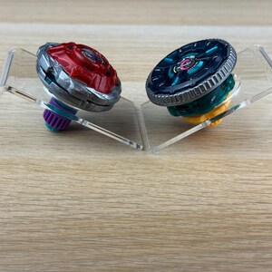 May include: Two spinning top toys displayed on clear acrylic stands. The left toy features a red metallic top with a silver rim and a purple base. The right toy has a dark teal metallic top with a silver rim and a yellow base. Both toys are circular.