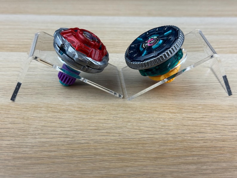 May include: Two spinning top toys displayed on clear acrylic stands. The left toy features a red metallic top with a silver rim and a purple base. The right toy has a dark teal metallic top with a silver rim and a yellow base. Both toys are circular.
