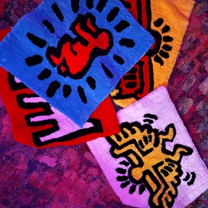 Op de afbeelding: Een verzameling kleurrijke, vierkante vloerkleden met opvallende grafische ontwerpen. De vloerkleden tonen iconische kunst in rood, blauw, geel en wit, met zwarte contouren. De ontwerpen omvatten een baby, een dansende figuur en andere abstracte vormen.