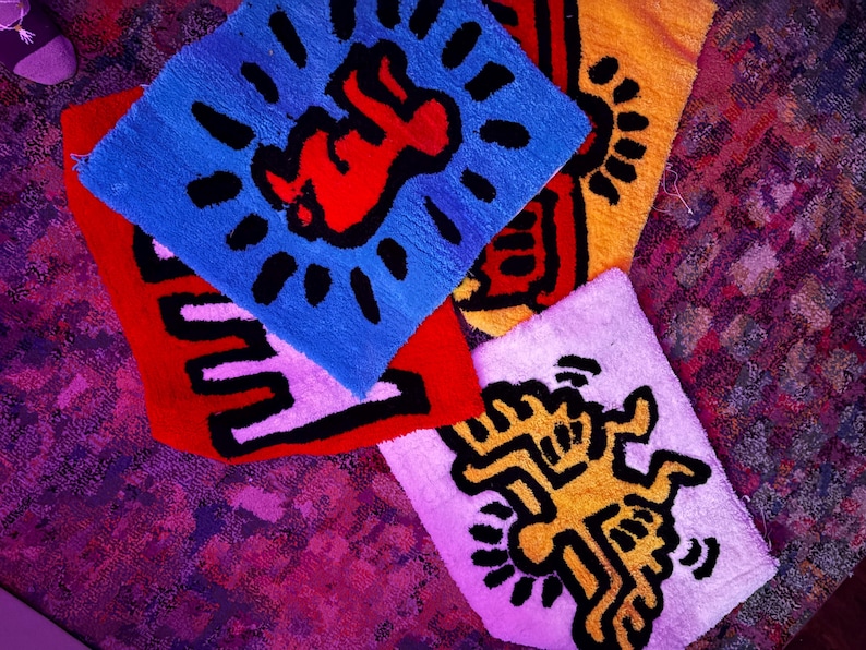 Op de afbeelding: Een verzameling kleurrijke, vierkante vloerkleden met opvallende grafische ontwerpen. De vloerkleden tonen iconische kunst in rood, blauw, geel en wit, met zwarte contouren. De ontwerpen omvatten een baby, een dansende figuur en andere abstracte vormen.