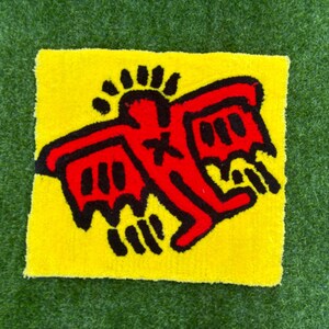 Op de afbeelding: Een vierkant, geel tapijt met een rode figuur en zwarte details, in de stijl van Keith Haring. De figuur heeft uitgestrekte armen en vleugels. Het tapijt ligt op een groene, grasachtige ondergrond.