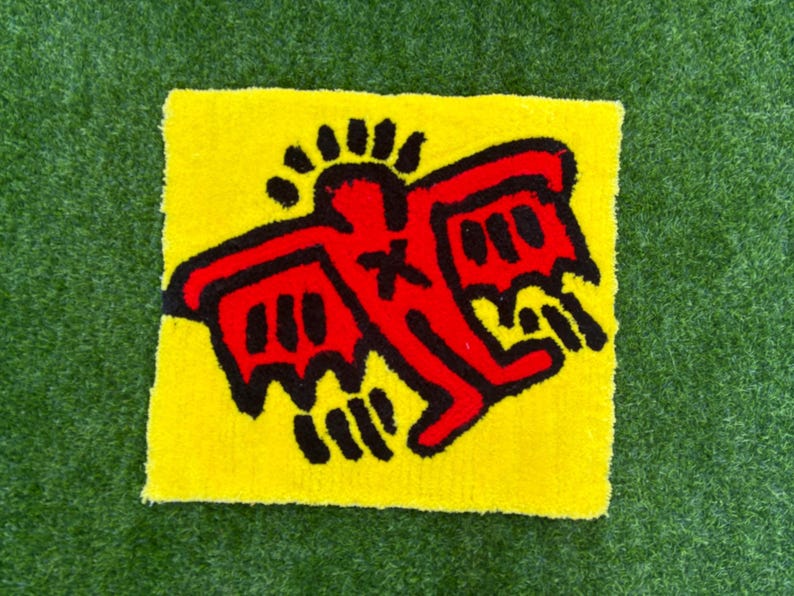 Op de afbeelding: Een vierkant, geel tapijt met een rode figuur en zwarte details, in de stijl van Keith Haring. De figuur heeft uitgestrekte armen en vleugels. Het tapijt ligt op een groene, grasachtige ondergrond.