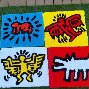 Op de afbeelding: Vier kleurrijke, vierkante tapijten met gedurfde grafische ontwerpen. Elk tapijt toont een ander Keith Haring-ge&iuml;nspireerd figuur in rood, geel en wit tegen blauwe, gele, witte en rode achtergronden. De tapijten liggen op een groene ondergrond.