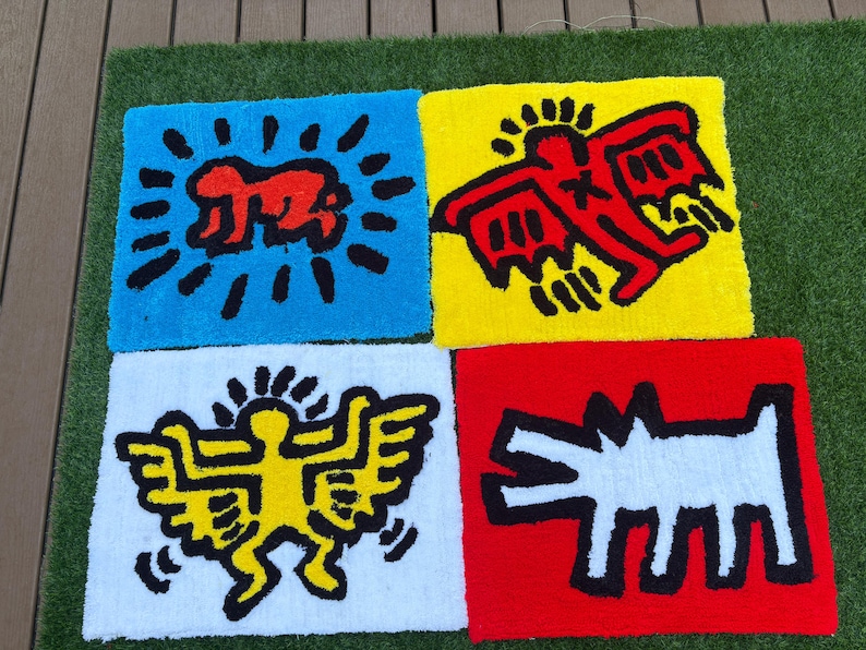 Op de afbeelding: Vier kleurrijke, vierkante tapijten met gedurfde grafische ontwerpen. Elk tapijt toont een ander Keith Haring-ge&iuml;nspireerd figuur in rood, geel en wit tegen blauwe, gele, witte en rode achtergronden. De tapijten liggen op een groene ondergrond.