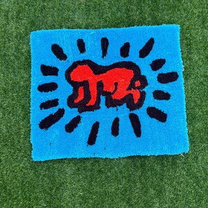 Op de afbeelding: Een vierkant, blauw tapijt met een rode figuur in het midden, omringd door zwarte stralende lijnen. Het tapijt heeft een zacht, getextureerd uiterlijk en het ontwerp doet denken aan pop-art.