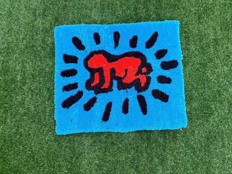 Op de afbeelding: Een vierkant, blauw tapijt met een rode figuur in het midden, omringd door zwarte stralende lijnen. Het tapijt heeft een zacht, getextureerd uiterlijk en het ontwerp doet denken aan pop-art.