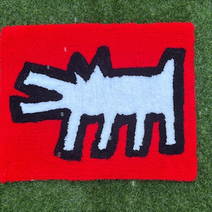 Op de afbeelding: Een rood tapijt met een witte, hondachtige figuur met zwarte contouren. Het tapijt heeft een rechthoekige vorm en ligt op een groene ondergrond. Het ontwerp is een opvallende, grafische weergave.