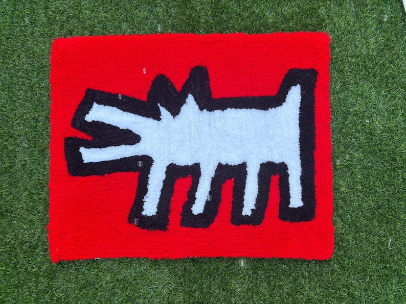 Op de afbeelding: Een rood tapijt met een witte, hondachtige figuur met zwarte contouren. Het tapijt heeft een rechthoekige vorm en ligt op een groene ondergrond. Het ontwerp is een opvallende, grafische weergave.