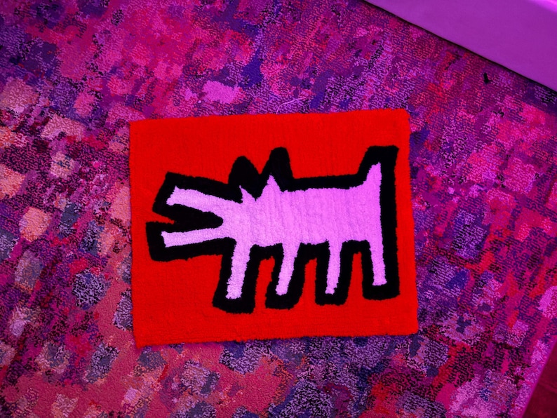Op de afbeelding: Een vierkant, rood tapijt met een witte, hondachtige figuur met zwarte contouren. Het tapijt heeft een textuur en ligt op een patroon, veelkleurig tapijt.