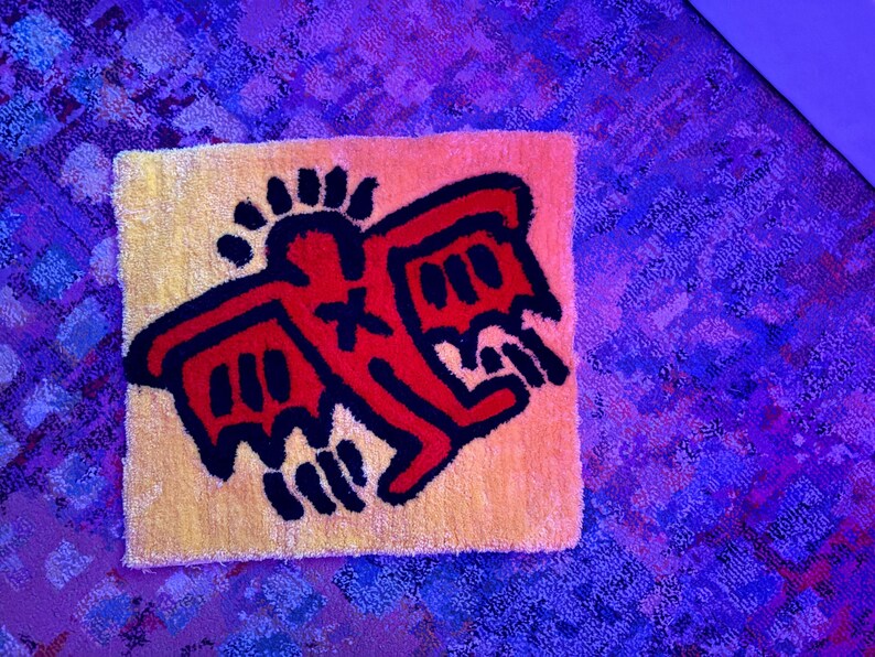Op de afbeelding: Een kleine, vierkante vloerkleed met een gele achtergrond en een rood-zwart grafisch ontwerp. Het ontwerp toont een gestileerde figuur met vleugels en een kruis. Het vloerkleed is waarschijnlijk gemaakt van een zacht, getextureerd materiaal.
