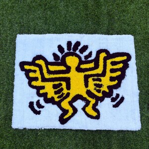 Op de afbeelding: Een rechthoekig wit tapijt met een gele figuur met zwarte contouren en vleugels, die doet denken aan de kunststijl van Keith Haring. De figuur is gecentreerd op het tapijt, tegen een groene gras achtergrond.