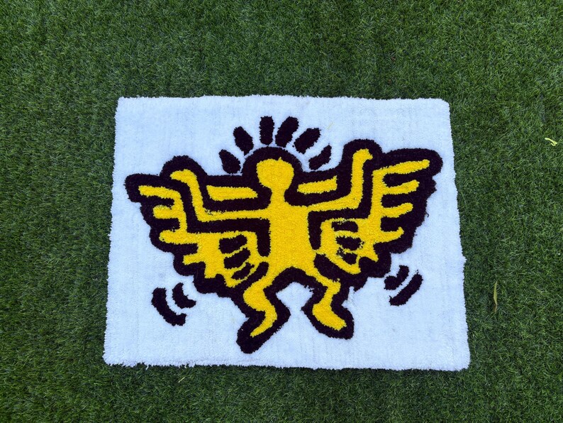 Op de afbeelding: Een rechthoekig wit tapijt met een gele figuur met zwarte contouren en vleugels, die doet denken aan de kunststijl van Keith Haring. De figuur is gecentreerd op het tapijt, tegen een groene gras achtergrond.