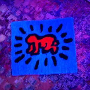 Op de afbeelding: Een rechthoekig blauw tapijt met een rode figuur en een zwart zonnestraalontwerp. Het tapijt heeft een zacht, getextureerd oppervlak. Het ontwerp is in pop-art stijl. Het tapijt is waarschijnlijk gemaakt van een pluche materiaal.
