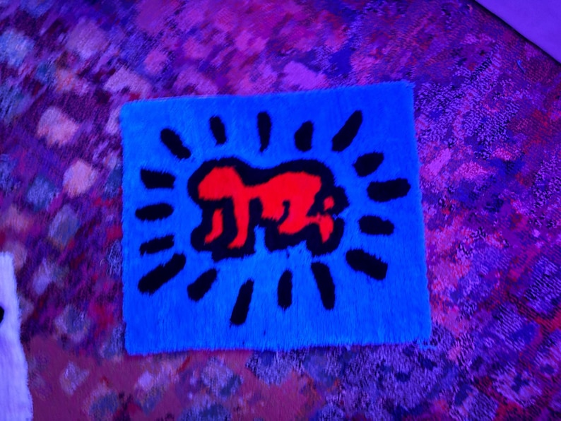 Op de afbeelding: Een rechthoekig blauw tapijt met een rode figuur en een zwart zonnestraalontwerp. Het tapijt heeft een zacht, getextureerd oppervlak. Het ontwerp is in pop-art stijl. Het tapijt is waarschijnlijk gemaakt van een pluche materiaal.