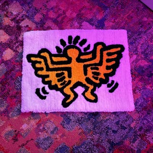 Op de afbeelding: Een wit rechthoekig tapijt met een opvallende oranje en zwarte afbeelding van een figuur met vleugels en een zon. Het ontwerp doet denken aan straatkunst.