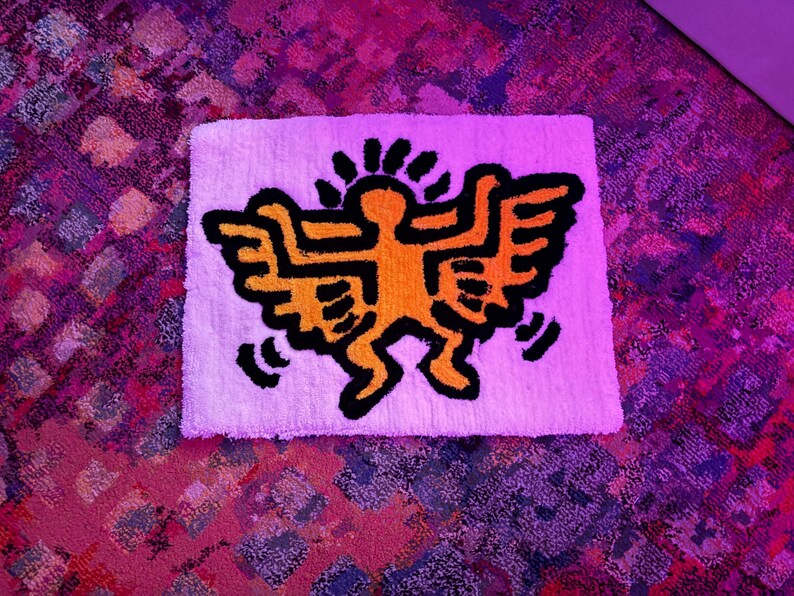 Op de afbeelding: Een wit rechthoekig tapijt met een opvallende oranje en zwarte afbeelding van een figuur met vleugels en een zon. Het ontwerp doet denken aan straatkunst.