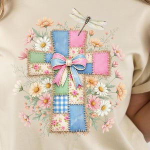 以下が含まれることがあります： ベージュのTシャツにカラフルな十字架のデザイン。十字架はピンク、青、緑、花柄のパッチワーク正方形でできており、ピンクと青のリボンで飾られています。トンボと花の要素が十字架を囲んでいます。