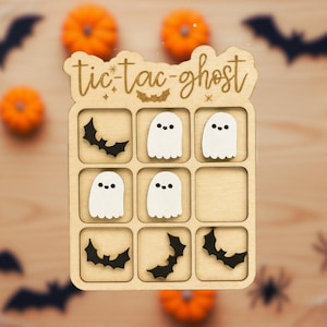 May include: A wooden tic-tac-toe game with a Halloween theme. The light brown game board has the words "tic-tac-ghost". The game pieces are white ghost shapes and black bat shapes. Pumpkins, bats, and spiders decorate the background.