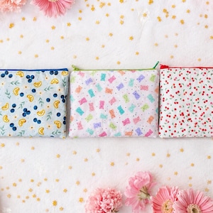 May include: Three rectangular fabric zipper pouches arranged side by side. The left pouch features a blue, yellow, and green fruit print. The center pouch displays a colorful gummy bear pattern. The right pouch shows a red cherry print.