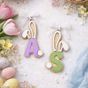 May include: Wooden Easter bunny-themed ornaments with the letters "A" and "S". The "A" is purple, and the "S" is green. Both have white bunny ears and a white bow. Decorative Easter eggs and flowers surround the ornaments.