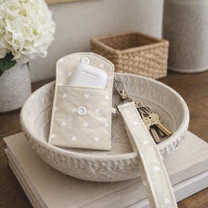 May include: A beige fabric pouch with white heart patterns, holding white earbuds, sits in a decorative bowl. A matching wrist strap with keys and a snap closure is next to the pouch. The items are on a stack of books.