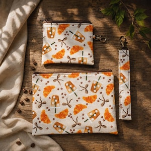 May include: Three cream-coloured pouches featuring a coffee and pastry design. The pattern includes orange croissants, iced coffee, coffee beans, and brown bows. One pouch has a zip and a keychain, another has a zip, and the third is a wristlet.