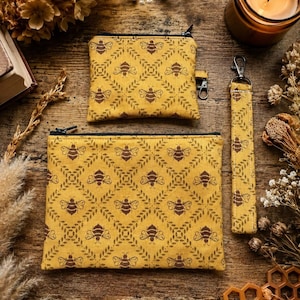 Peut inclure: Ensemble d'accessoires en tissu jaune avec un motif répété d'abeilles et de feuilles marron. L'ensemble comprend une petite pochette zippée, une plus grande pochette zippée et un porte-clés. Sur une surface en bois rustique.