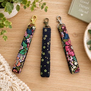May include: Three fabric wristlet keychains with metal lobster clasps. Two wristlets feature a colourful floral pattern with pink, green, and purple shapes. The centre wristlet is dark blue with a small yellow flower pattern and a bronze-tone clasp.