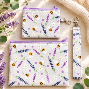 May include: A set of three fabric accessories: a small zipped pouch, a larger zipped pouch, and a wristlet. The light grey fabric is patterned with bees, lavender sprigs, and small white flowers. The pouches have purple zips.