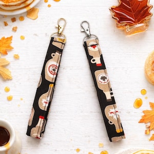 May include: Two black fabric keychains with a maple syrup bottle pattern, gold and silver clasps. The image also shows pancakes with butter, maple syrup, and a maple leaf-shaped bottle.