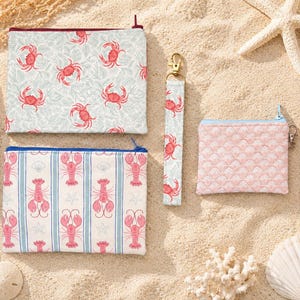 May include: A collection of beach-themed accessories, including zippered pouches and wristlet keychains. The pouches feature patterns of crabs, lobsters, and a seashell design. The color palette includes pink, blue, and white, evoking a coastal aesthetic.