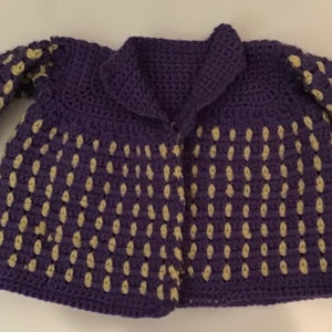 May include: A purple crocheted cardigan with a yellow dot pattern. The cardigan has a collar and long sleeves.
