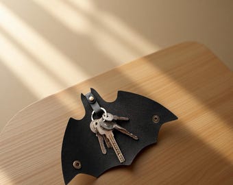 May include: A black leather key organizer shaped like a bat silhouette. The item features a central metal snap closure and a metal ring holding a set of silver-toned keys. The design includes two additional metal snaps on the wings for folding.