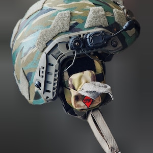 May include: A tactical helmet featuring a green, brown, and black camouflage pattern with beige velcro patches. It includes a dark gray side rail system, a black communication mount, and a small red diamond-shaped patch with black lettering.