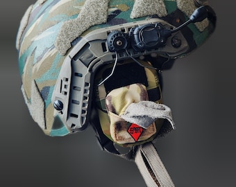 May include: A camouflage helmet with a black and gray tactical headset and a small pouch attached to the side. The pouch has a red diamond-shaped patch with the word "FAST" written on it.
