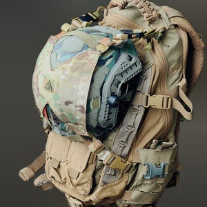 May include: A tan tactical backpack with multiple pockets and straps. A camouflage helmet is secured to the front of the pack. The helmet has a dark gray face shield and is partially covered by a camouflage cover. The backpack has multiple buckles and straps.