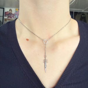 May include: A silver-colored necklace featuring a small dagger pendant. The pendant hangs from a delicate chain that connects to a circular ring. The necklace is worn against a dark blue top.