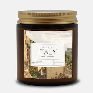 May include: A brown glass candle jar with a gold lid. The label features a photo of an Italian village and the text "Smells Like Italy Sage & Cypress". The candle is made with coconut soy wax.