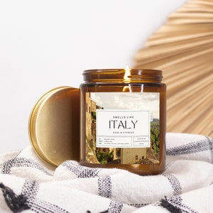 May include: A brown glass candle jar with a gold lid. The label says "Smells Like Italy" and "Sage & Cypress". The label also features a photo of an Italian village.